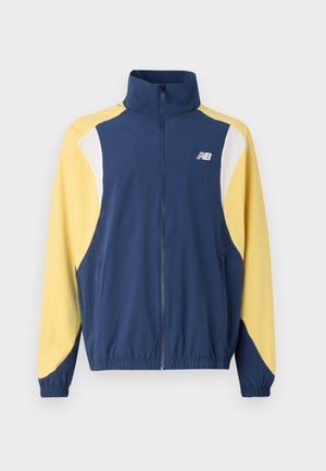 Marineblauwe en gele ritsjack met hoge kraag, met een kleurblokontwerp en elastische manchetten; de stof lijkt glad en sportief.