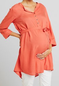 Túnica de maternidad coral con escote redondo, detalles de botones y cintura anudada. Presenta mangas tres cuartos y un dobladillo curvado.