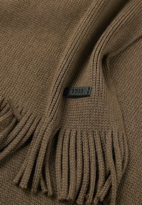 Écharpe en tricot brun avec des bords à franges, fabriquée en tissu texturé. Elle présente une petite étiquette de logo noire attachée au matériau.