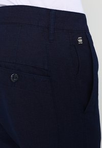 Marineblauwe broek van gestructureerde stof met een knoopsluiting en een klein logo detail vlakbij de tailleband. Voorzien van een zijzak.