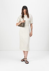 Valkoinen pellavamekko puffi-hihoilla, istuvalla vyötäröllä ja vyöllä. Malli kantaa pientä harmaata käsilaukkua ja käyttää mustia remmikenkä.