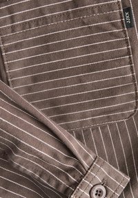 Brown striped fabric with white lines; features a button on the cuff and a patch pocket; smooth texture, woven design.