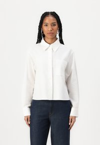 Camicia bianca a maniche lunghe con colletto a punta, tasca sul petto e orlo corto. Indossata con pantaloni di jeans blu scuro.