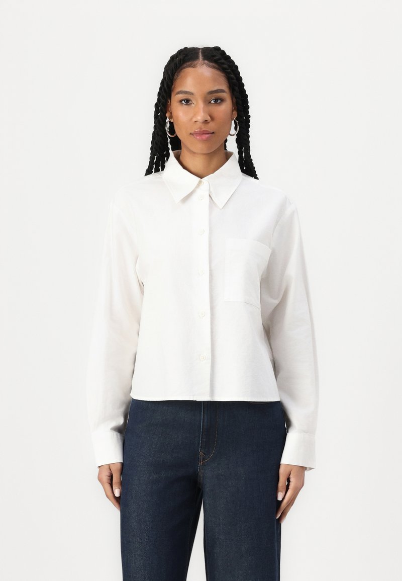 Camicia bianca a maniche lunghe con colletto a punta, tasca sul petto e orlo corto. Indossata con pantaloni di jeans blu scuro.
