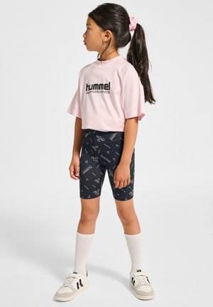 Jong meisje met lang donker haar in een paardenstaart, draagt een roze Hummel Performance-shirt, zwarte korte broek met patroon, witte sokken en sneakers, staand zijwaarts.