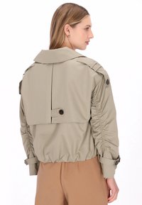 Beige, leichte Jacke mit tailliertem Bund, großem Kragen und Knopfakzenten. Verfügt über geraffte Ärmel und ein markantes Rückendesign.
