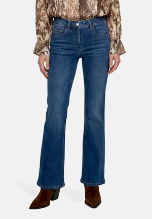 Femme portant un jean bootcut bleu, des bottines en daim marron à talons et un chemisier à manches longues à motifs avec des poignets évasés.