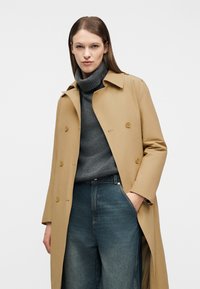 Γυναίκα που φοράει ένα μπεζ παλτό με διπλή σειρά κουμπιών πάνω από γκρι ζιβάγκο πουλόβερ και μπλε τζιν με ψηλή μέση, στέκεται με το ένα χέρι στην τσέπη.