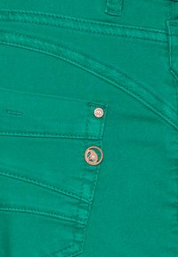Tecido de denim verde com uma textura suave, apresentando bolsos angulados e um botão em rosa dourado com um detalhe em design floral.