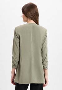 Olijfgroene blouse met lange mouwen en een relaxte pasvorm. Heeft een eenvoudig ontwerp, een soepele textuur en gegatherde details aan de mouwen.