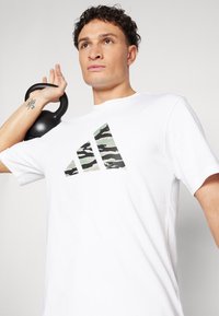 Bijela majica kratkih rukava s uzorkom vojničke kamuflaže u obliku tri pruge preko prsa. Muškarac drži crnu kettlebell.