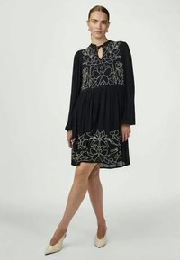 Črna obleka z dolgimi rokavi, ki ima belo cvetlično vezenje, vratni trak in razširjen kroj. Gladka tkanina z mehkimi teksturami.