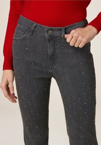 Personne portant un jean gris foncé avec de petites clous en argent et un haut à manches longues rouge, main ajustant la taille sur le côté droit.