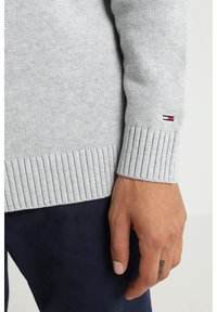 Primo piano di una mano appoggiata al polsino di un maglione grigio a maglia con un piccolo logo rosso, bianco e blu, abbinato a pantaloni blu scuro.