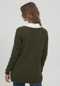 Pletený svetr v tmavě zelené barvě s texturovaným vzorem, dlouhými rukávy a kulatým výstřihem, přetažený přes bílou košilovou košili.