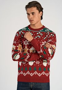 Punainen neulottu joulupaita, jossa on karkkikeppejä, piparkakkuja, lumihiutaleita, puita ja joulupukki-aiheita. Kohokuvioitu kaula-aukko ja hihansuut.