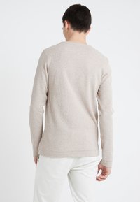 Man i en beige långärmad tröja och vita byxor, sedd från baksidan mot en enfärgad vit bakgrund.