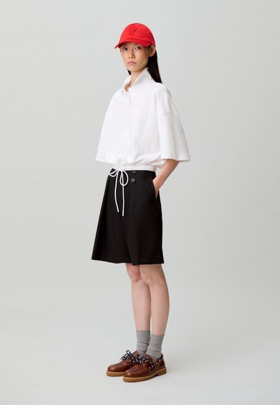 Polo blanc oversize avec taille à cordon, short noir avec boutons, chaussettes grises et chaussures marron avec lacets décoratifs. Casquette rouge.