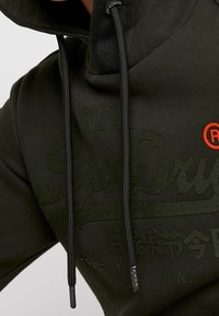 Sweat à capuche vert foncé avec cordons noirs, texte embossé « SuperDry », caractères japonais et un petit symbole rouge de marque déposée sur la poitrine.