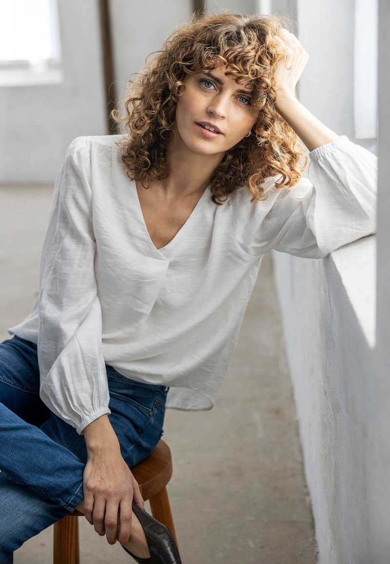 Femme aux cheveux bouclés en blouse blanche et jean bleu, assise sur un tabouret en bois, appuyée sur un rebord de fenêtre blanc, regardant calmement devant elle.