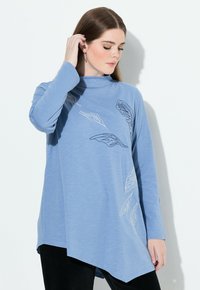 Ljust blå långärmad topp med en ståkrage, med diagonal fåll och vita bladmotiv broderade på framsidan. Mjuk tygkvalitet.