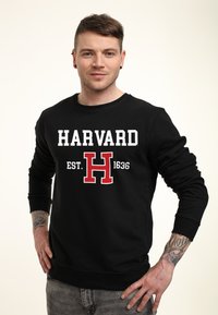 Felpa nera con la scritta "HARVARD" in bianco e rosso, con "EST. 1636" e una lettera "H" rossa. Vestibilità comoda con polsini a costine.