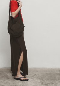 Haut rouge à manches courtes, jupe maxi marron avec fente sur le côté, sac à épaule en daim marron et tongs noires sur une surface en béton.