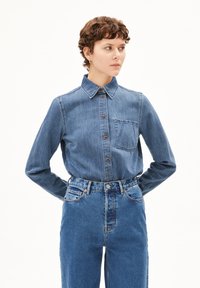 Denim overhemd met knopen en kraag, blauwe stof, voorzak en bruine knopen. Gecombineerd met hoog-waist blauwe jeans.