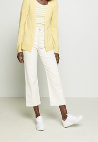 Letvægts gul cardigan over en hvid og stribet skjorte, parret med hvide brede jeans og hvide sneakers. Glat tekstur.