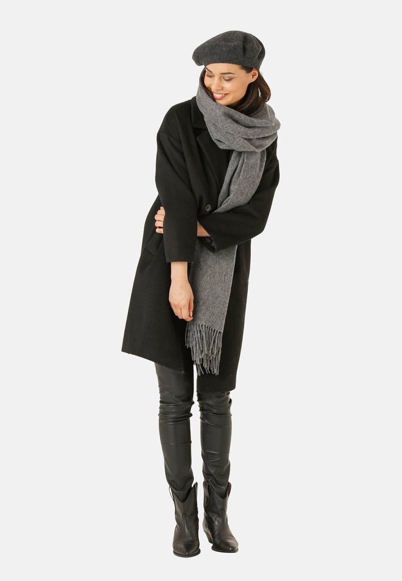 Sort uldfrakke, grå uldsjaal med frynser, sorte læderbukser og en grå beret, hvilket skaber et lagdelt, monokromatisk look.