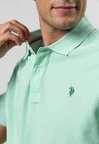 Világos zöld pólóing texturált mintával, két gombos gallérral és hímzett logóval a bal mellen.