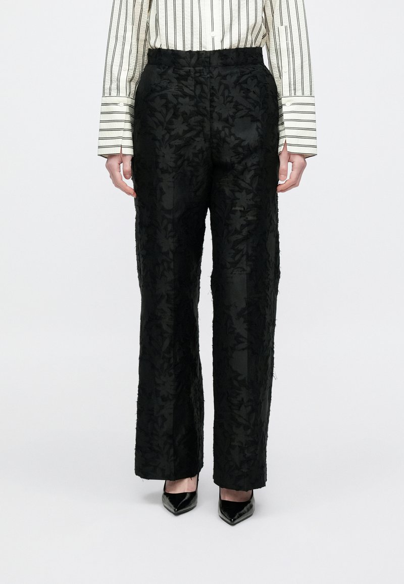 Calças pretas com padrão floral texturizado, design de cintura alta e corte de perna reta. Combinadas com uma camisa de botões às riscas e saltos altos.