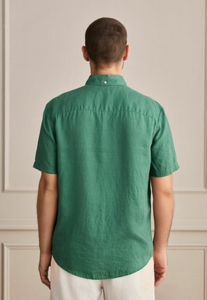 Mees seisab seljaga vaataja poole, tal on seljas lühikeste varrukatega roheline särk ja valged püksid heleda beežika seinaga, millel on vormistus.