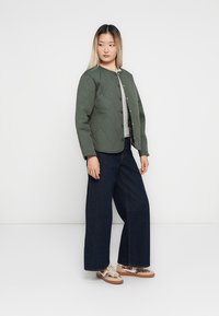 Veste verte matelassée avec un col rond et des boutons-pression, associée à une chemise grise, un jean large foncé et des baskets à motifs.