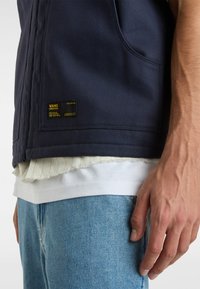 Gilet Navy Vans con una patch logo visibile, indossato sopra una maglietta bianca, abbinato a jeans in denim azzurri chiari. Tessuto texturizzato con un dettaglio della tasca.