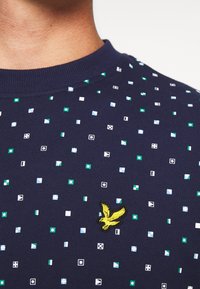 Felpa blu navy con colletto tondo, caratterizzata da motivi geometrici colorati e un logo di un uccello giallo ricamato sul petto.