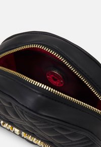 Black quilted leather pouch with gold zipper partially open, revealing red interior and circular logo tag inside.