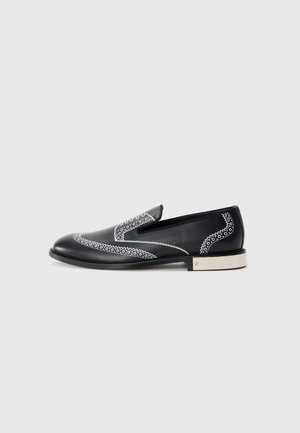 Melna ādas loafers kurpes ar baltu dekoratīvo šuvumu un metāla akcentu uz papēža, ar gludu tekstūru un plūstošu siluetu.