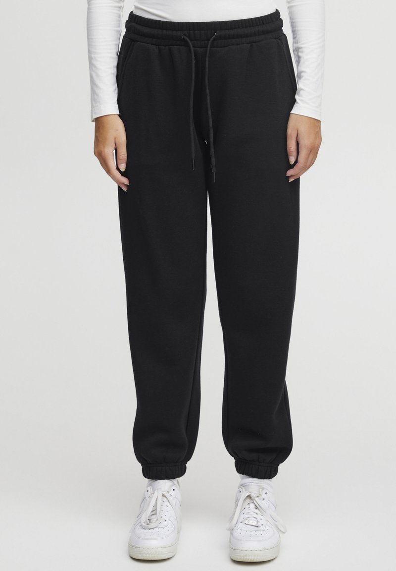 Svarta bomullsjoggare med elastisk midja och dragsko, försedd med ribbade ärmslut och sidofickor, i kombination med vita sneakers.