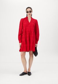Röd ögla klänning med långa ärmar, V-ringning, flerlagat fåll och vågiga kanter. Åtföljd av svarta accessoarer och platta skor.
