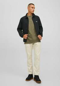 Giacca leggera nera con zip, t-shirt verde oliva con testo, pantaloni color crema e scarpe nere con suole marroni.