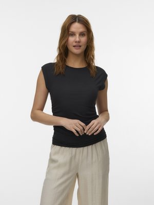 Γυναίκα με κυματιστά καστανά μαλλιά, που φοράει μια εφαρμοστή μαύρη αμάνικη μπλούζα και φαρδιά μπεζ παντελόνια, στέκεται μπροστά σε έναν απλό λευκό φόντο.