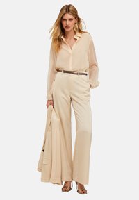 Blusa de seda beige con botones en la parte frontal y puños con volantes, combinada con pantalones anchos beige y una chaqueta a juego colgada en un brazo. Calzado con acabados en dorado.