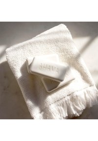 Tre vita AHAVA-tvålbarer med präglade bokstäver ligger på en strukturerad vit handduk med fransar. Mjuk belysning framhäver scenen.