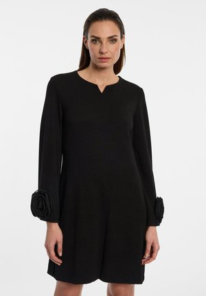 Vrouw die een zwarte jurk met lange mouwen draagt, met een kleine V-hals en grote stoffen bloemendetails op elke mouw bij de polsen.
