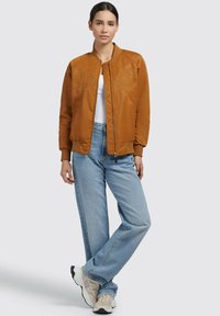 Giacca bomber marrone con ricami floreali, chiusura con zip, polsini e orlo a coste, abbinata a jeans azzurri e sneakers beige su uno sfondo grigio.