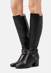 Bottes noires en cuir montant jusqu'aux genoux avec une finition texturée, une bride à la cheville ornée d'accents logo dorés, et un talon bloc. Fermeture éclair sur le côté.
