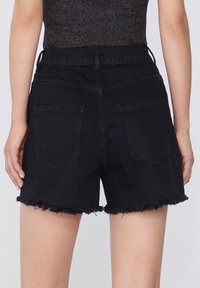 Bermudas de mezclilla negras con un dobladillo deshilachado, que cuentan con dos bolsillos traseros y una cintura de altura media. La textura es suave con un acabado mate.