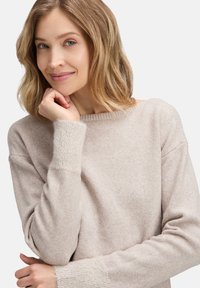 Jasno beżowy sweter o miękkiej fakturze, szerokim dekolcie i teksturowanych, prążkowanych mankietach. Projekt ma luźny krój i obniżone ramiona.