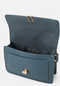 Borsa a tracolla sintetica turchese con hardware dorato, dotata di chiusura a patta, tasca frontale e texture liscia. L'interno presenta numerosi scomparti.